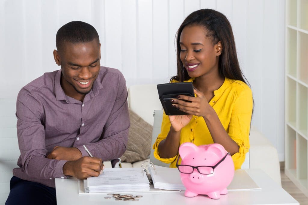 Happy Young Couple Calculating Bill With Coins And Piggybank On Desk - Budgeting Tips