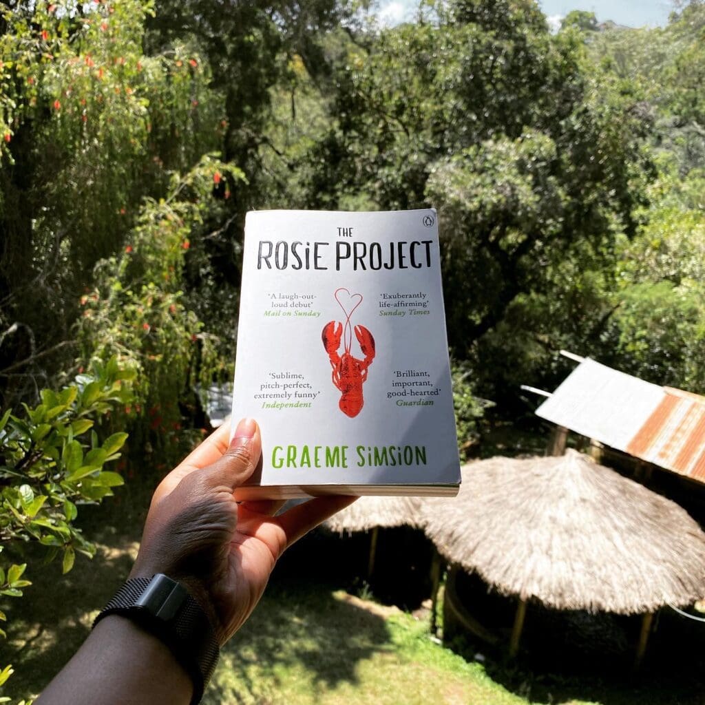 Holding a copy of the Rosie Project with houses in the background