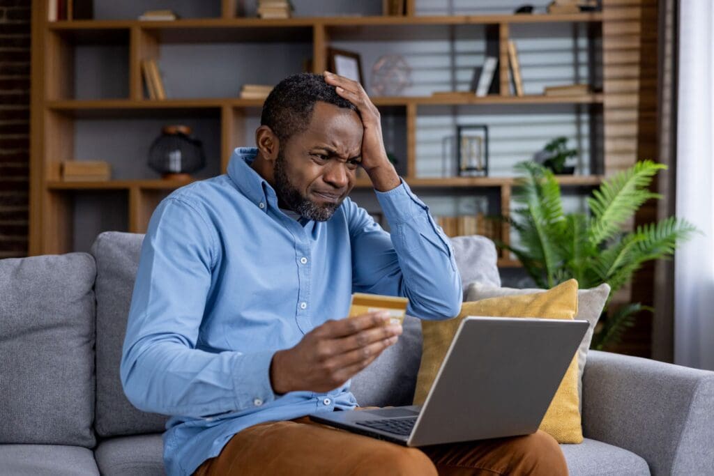 Worried african american man with credit card and laptop experiencing financial stress at home