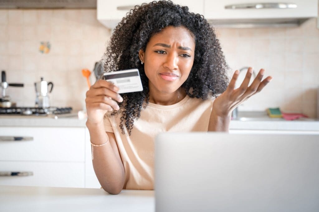 A lady holding a credit card, looking at her laptop screen and very stressed about money. An emergency fund can help meet such unexpected expenses. 