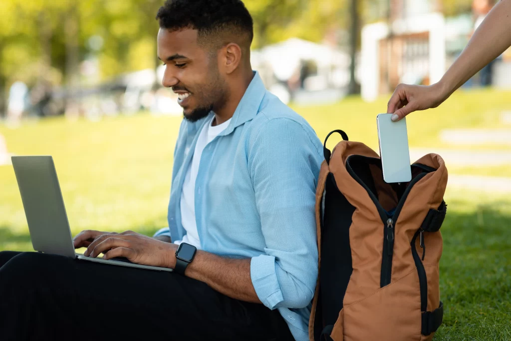 A mans hand pick pocketing a smartphone from a a young man who is sitting smiling at their laptop with their backpack wide open