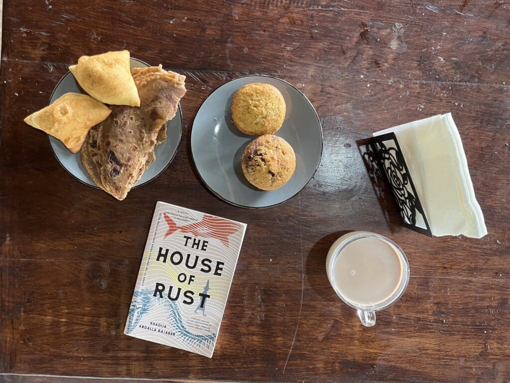 The House of Rust by Khadija Abdalla Bajaber novel and Snacks, cupcakes, mandazi and chapati with a cup of cake and serviettes on a table