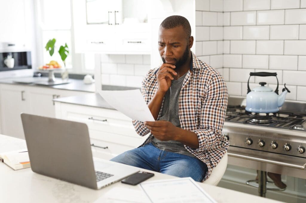 Serious African american man sitting in kitchen working looking at paperwork and using laptop