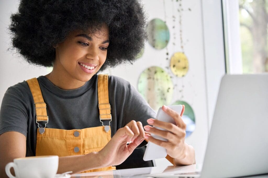 Afro American young woman in cafe holding phone with laptop shopping online