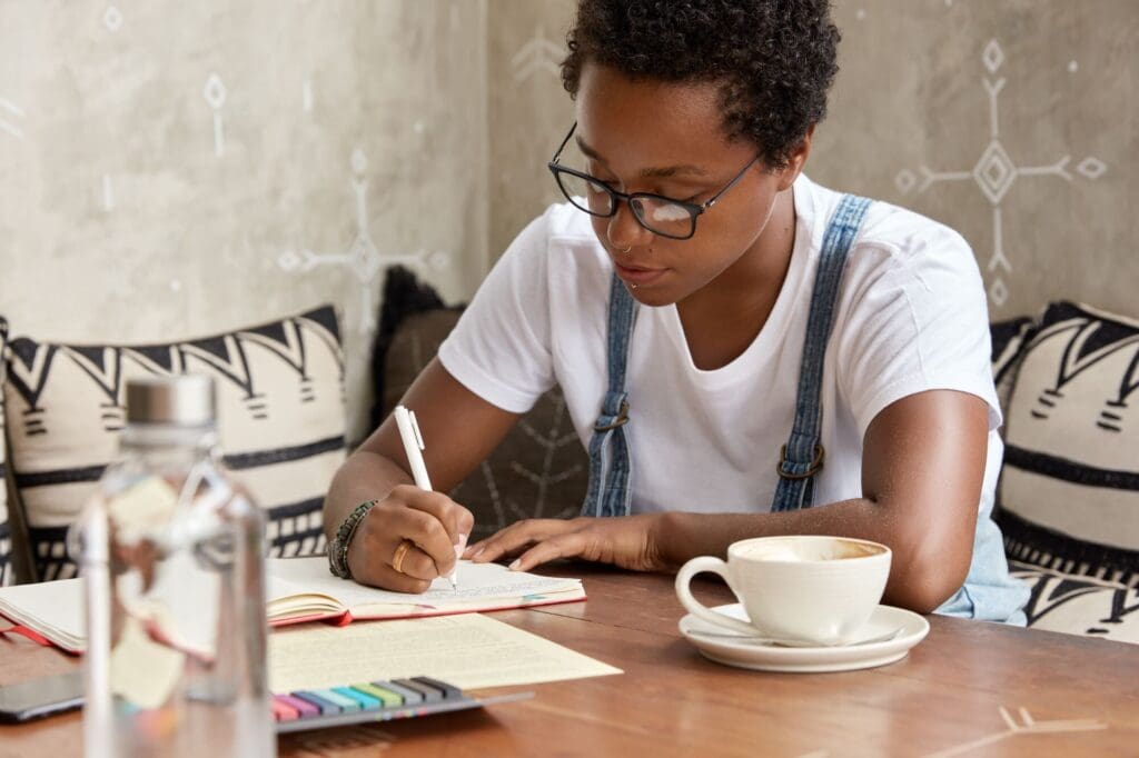 Young African woman in glasses writing in a notebook with a cup of coffee or tea