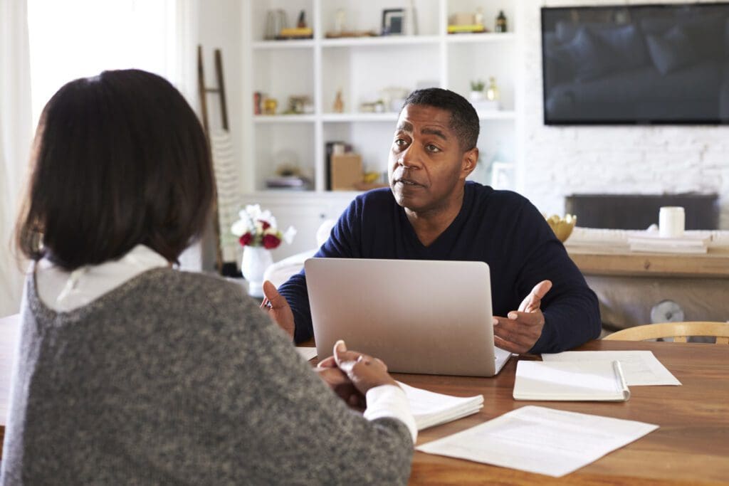 Millennial man with laptop computer giving financial advice to a woman sitting at the table in her dining room, selective focus