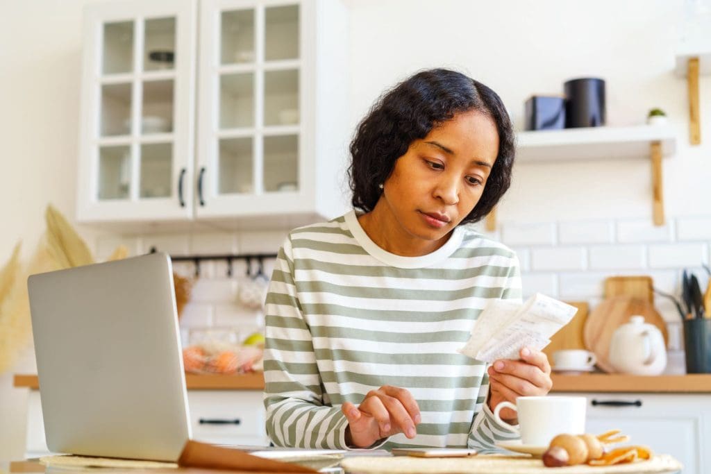 Young African woman sitting behind a kitchen counter with an open laptop and looking at a receipt and entering data into her phone