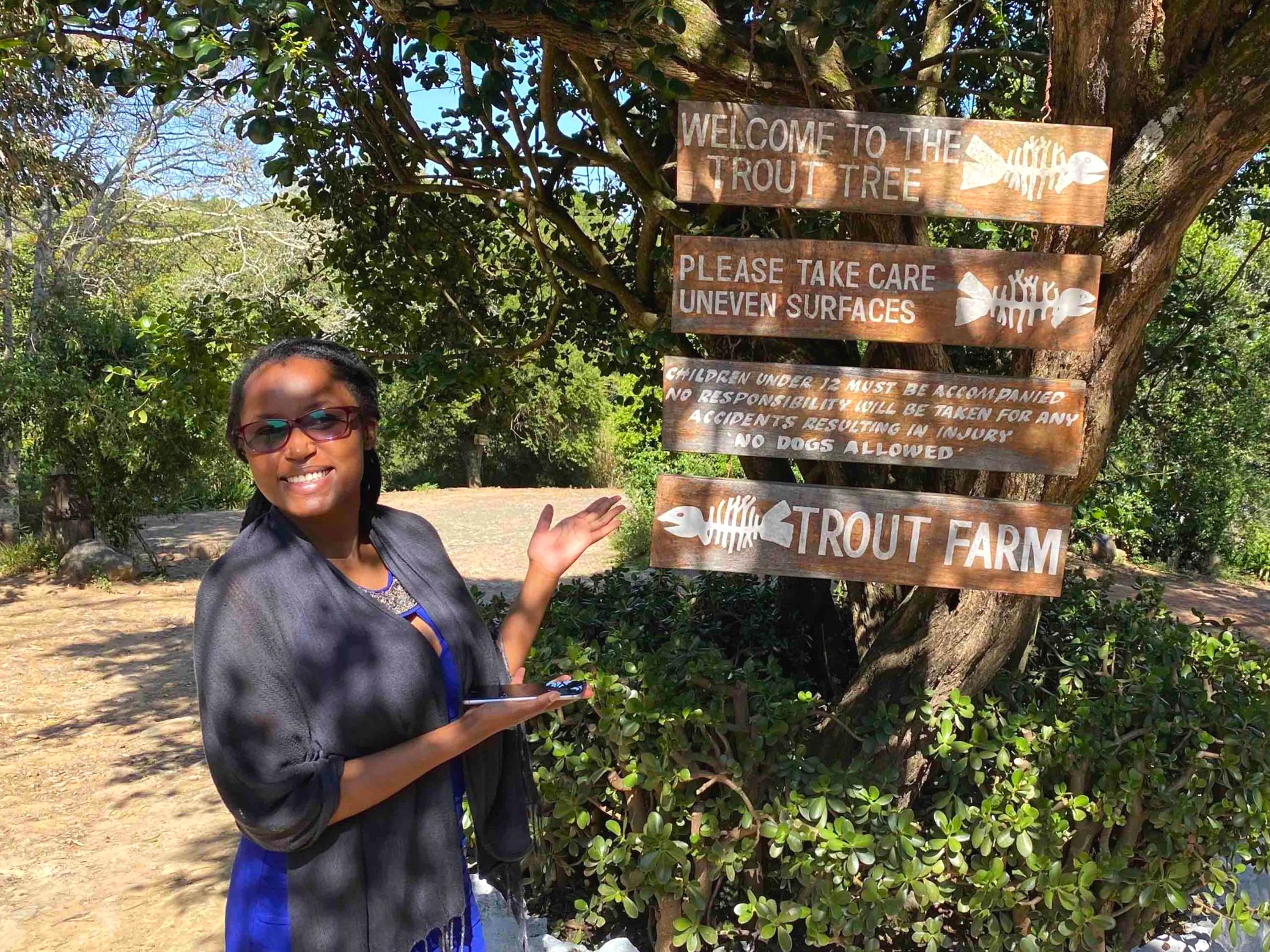 A young lady smiling and pointing at an entrance signage of The Trout Tree Restaurant in Nanyuki