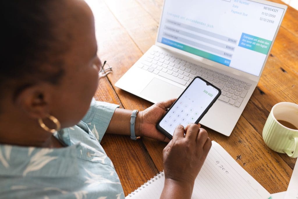 Busy senior African woman with documents on table calculating on smartphone at home
