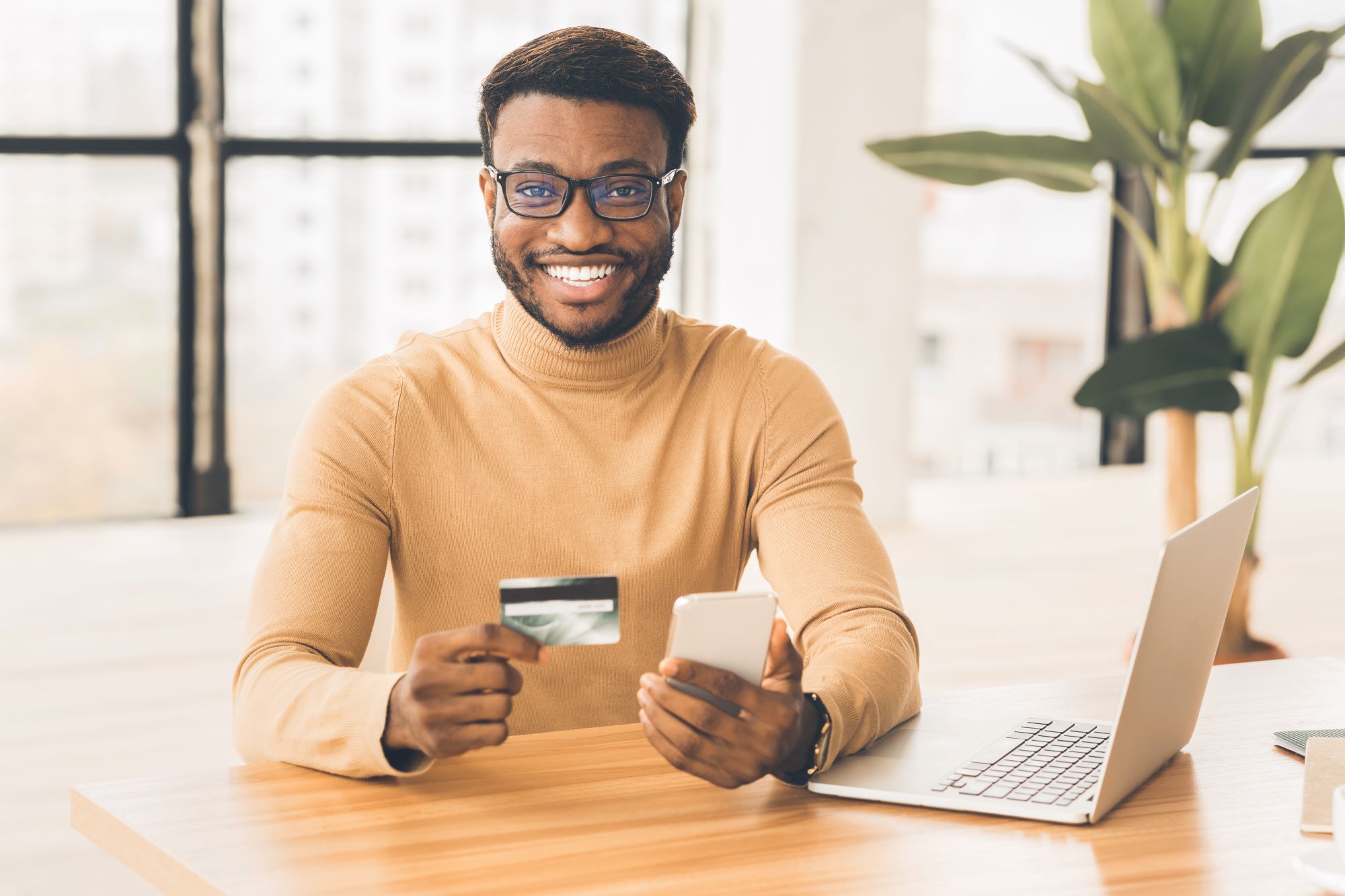 A young man smiling, holding smart phone and credit card trying internet banking