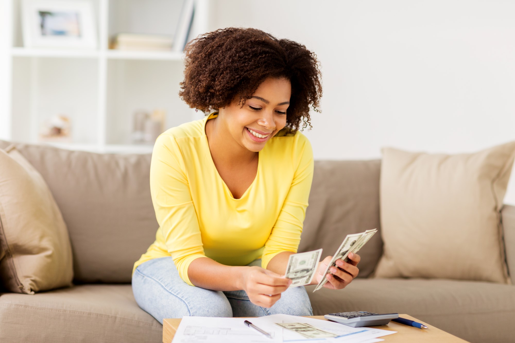 Financial wellness challenges; A young Black lady sitting on a couch, smiling and holding money