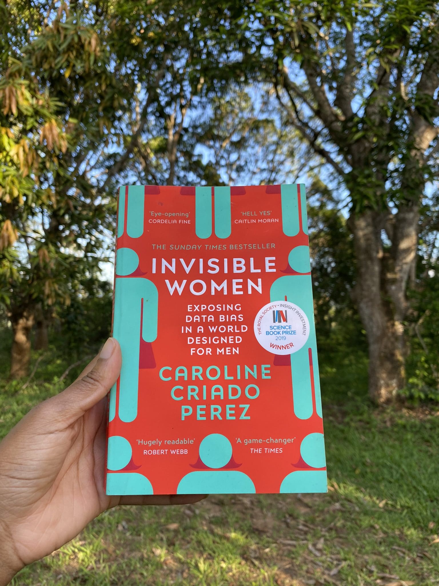 A Black lady's hand holding a copy of Invisible Women Data Bias in a World Designed for Men by Caroline Criado Pérez with a view of trees in the background