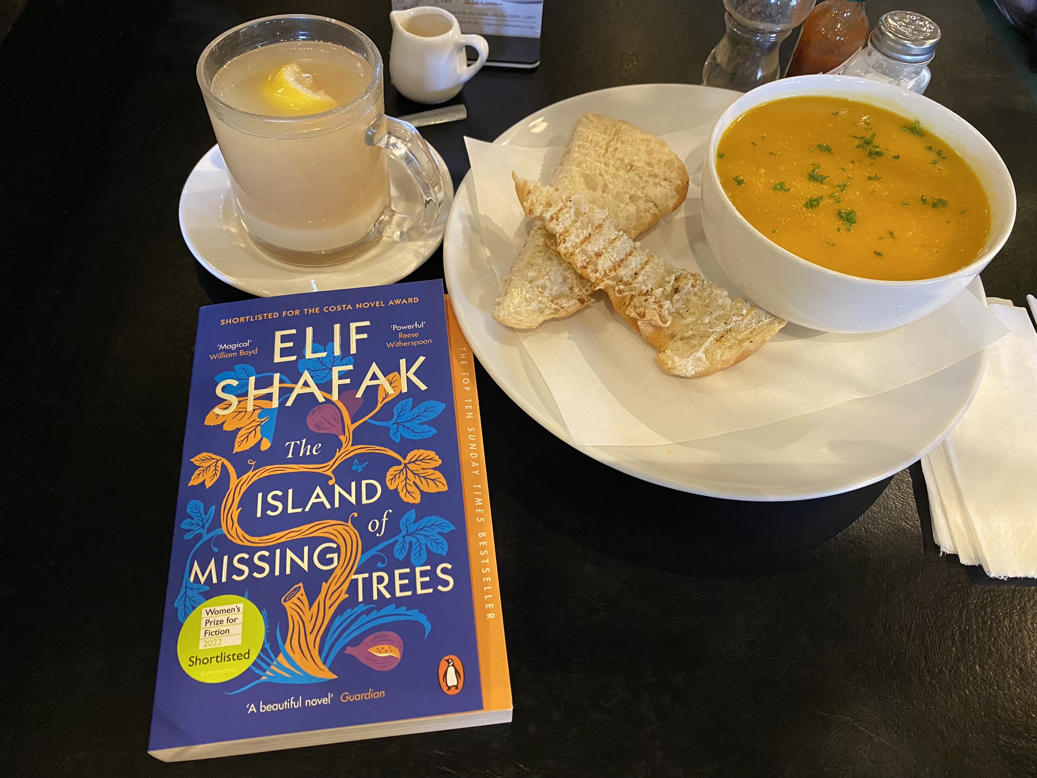 The Island of Missing Trees by Elif Shafak on a cafe's table with a bowl of soup, a side plate with ginger bread, and a glass of hot lemon and ginger tea
