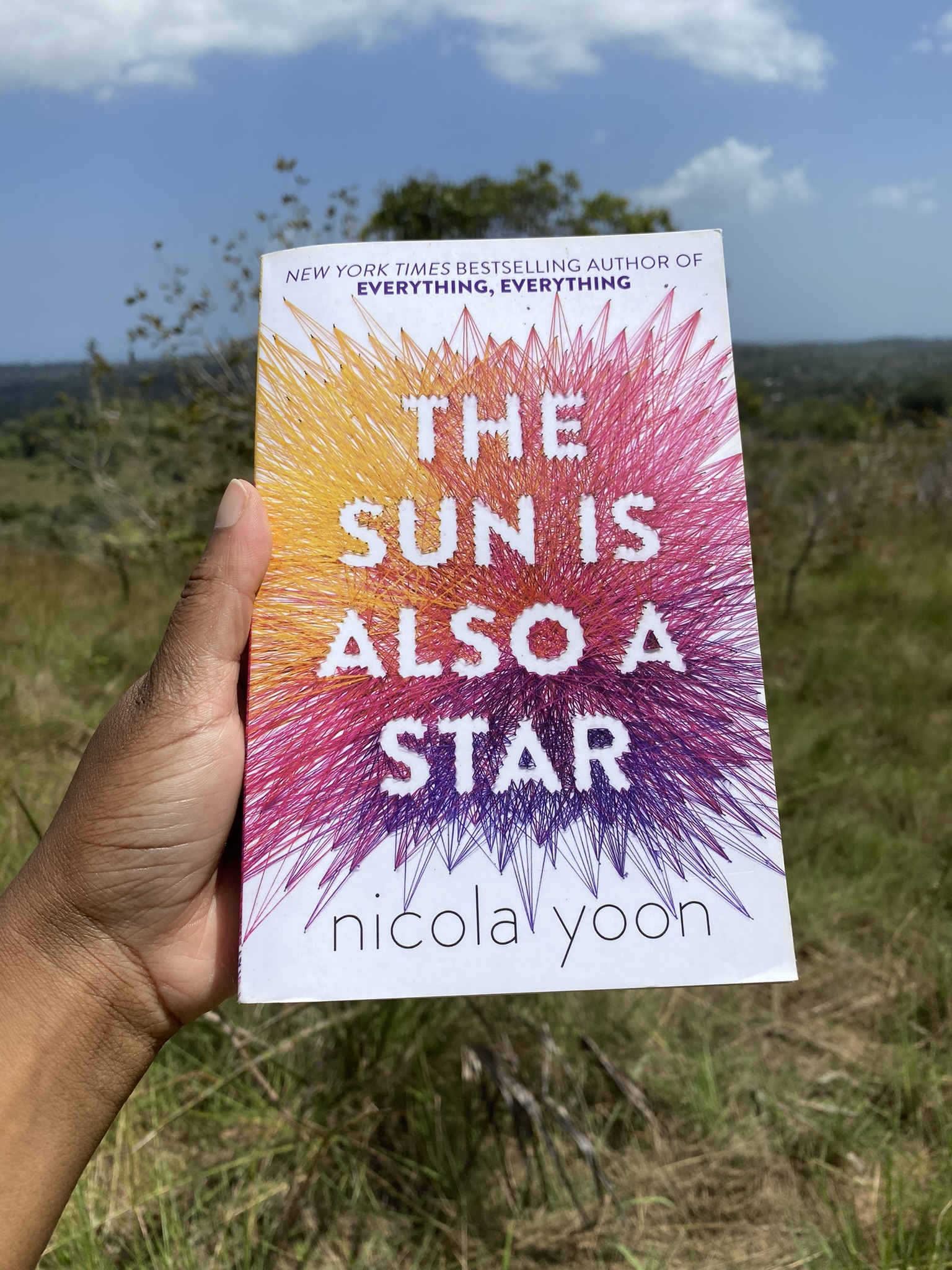 A Black lady's hand holding a copy of The Sun Is Also a Star by Nicola Yoon on a scenic view background