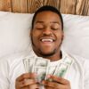 Happy African Man Holding Money Counting Cash Lying In Bed- enjoying returns from Fixed Income Funds